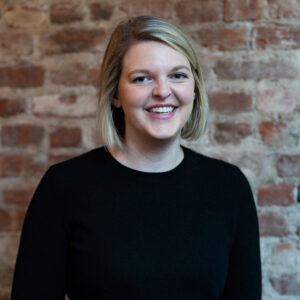 Portrait of a smiling woman with short blonde hair in a black sweater, standing against a brick wall.
