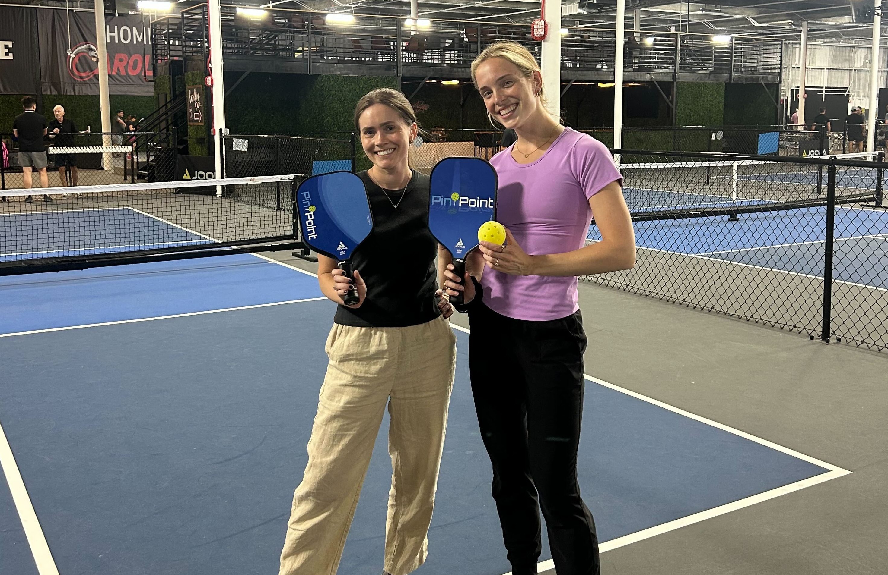 Two women stand on a blue indoor tennis/ping-pong court, smiling and holding blue PingPoint paddles with a yellow ball in one hand.