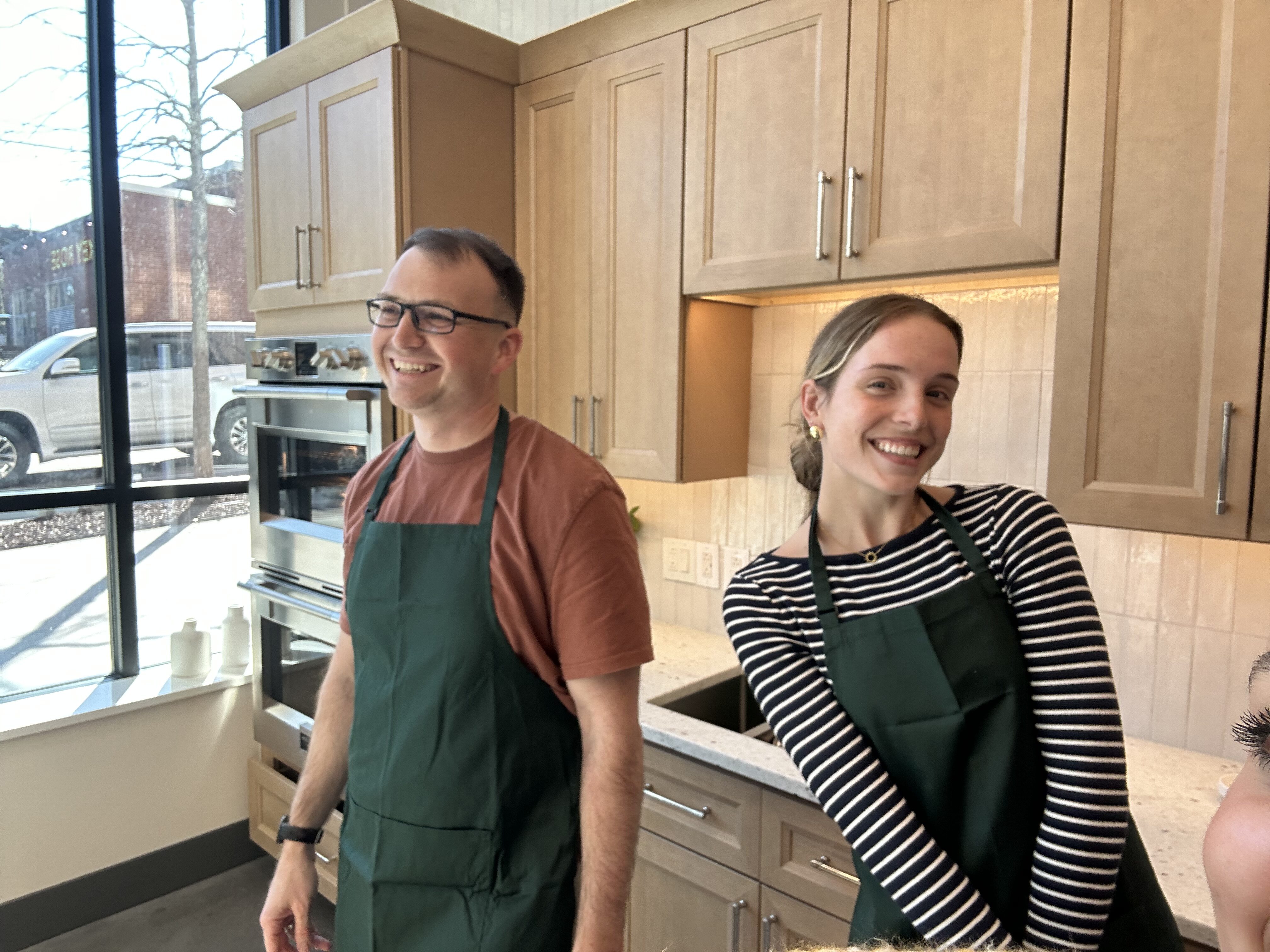 Two coworkers wearing green aprons smile at the camera in a bright kitchen showroom with wooden cabinets behind them.