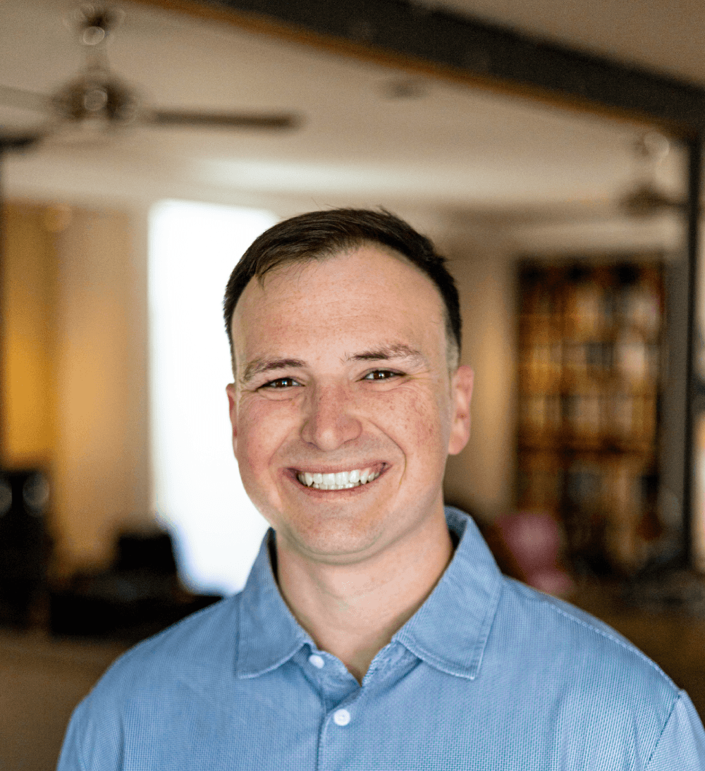 Smiling man in a light blue button-down shirt indoors with a blurred office background.