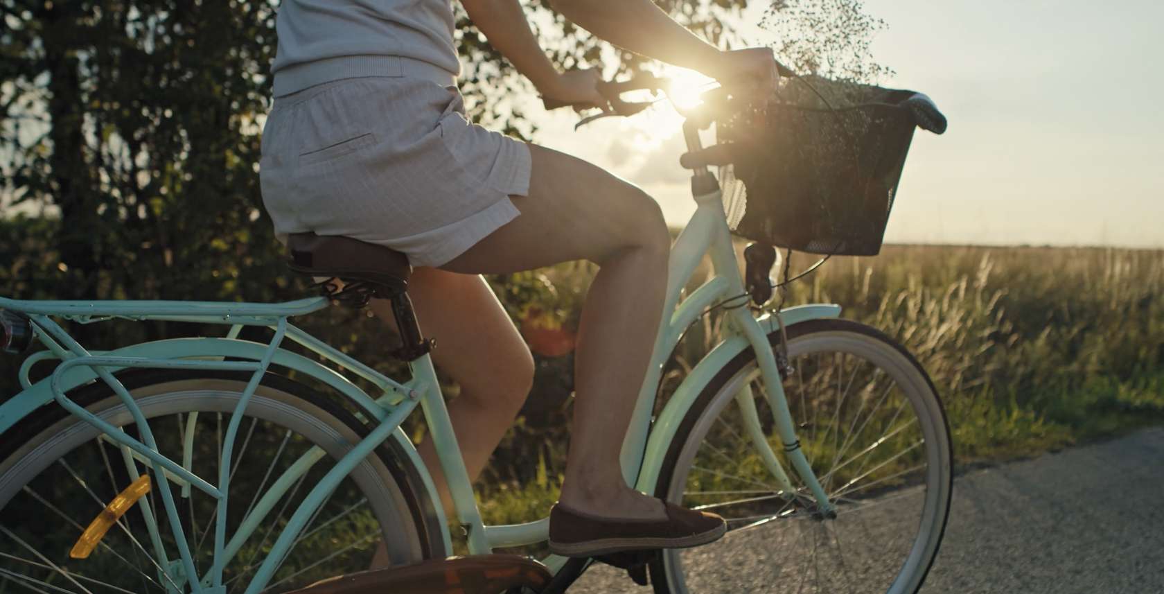 Person riding a light blue bicycle on a sunlit rural road, basket on the front, wearing shorts and loafers with the sun behind them.