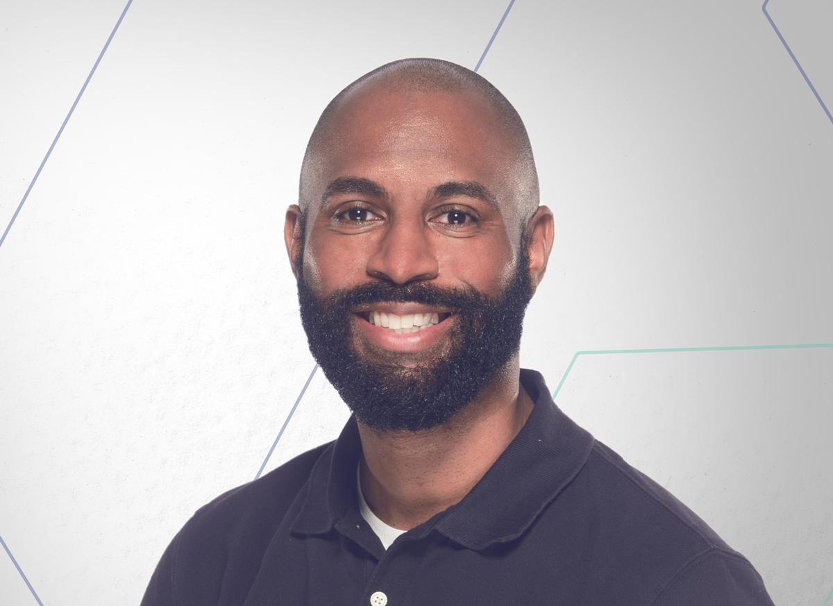 Smiling man with a full beard wearing a dark polo shirt, against a light geometric-patterned background.