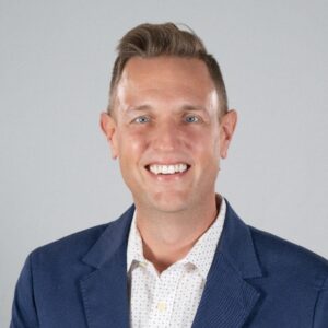 Smiling man in a navy blazer and white patterned shirt, portrait against a light gray background.