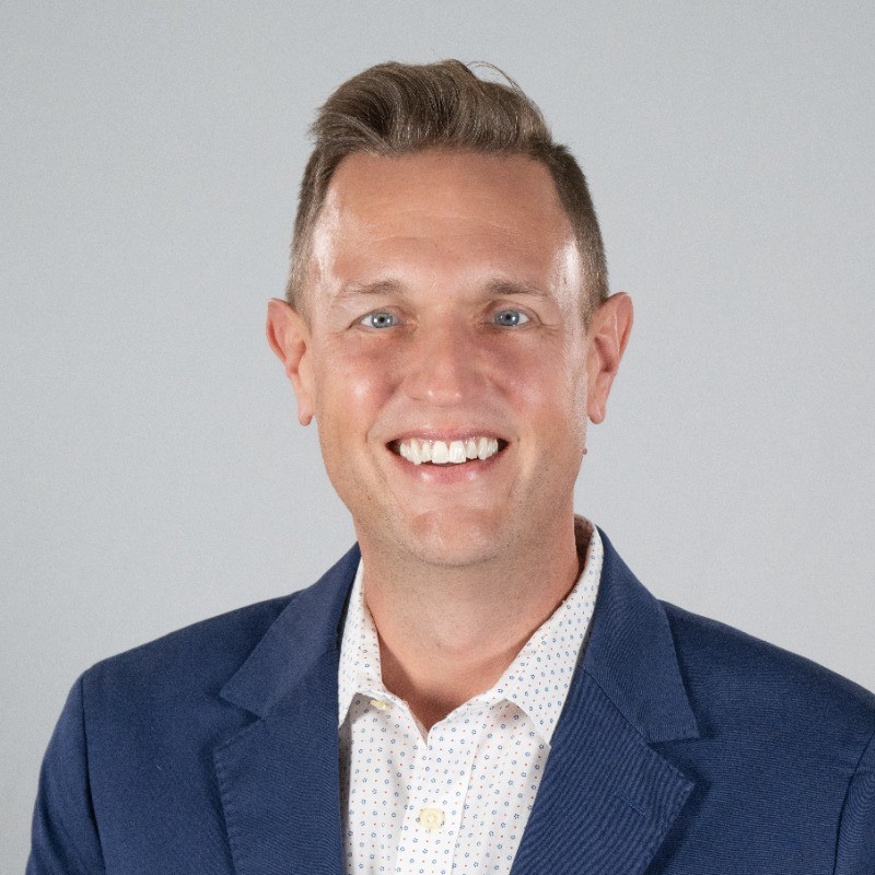 Smiling man in a navy blazer and white patterned shirt, portrait against a light gray background.