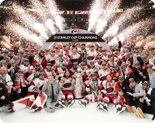 The 2026 Stanley Cup champions hockey team posing with the Stanley Cup on the ice as fireworks explode overhead.