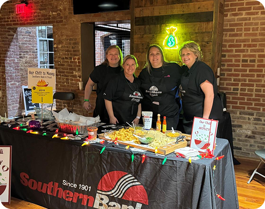 Four volunteers behind a Southern Bank fundraiser booth with chili and toppings, set in a brick-walled venue; a neon dollar sign glows on the wall.