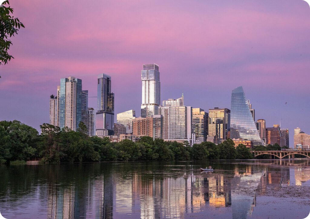 City skyline at sunset with glass skyscrapers reflecting in a calm river, trees along the bank, and a small boat on the water.