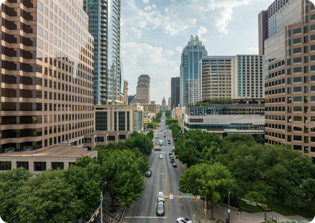 A wide city street lined with tall office buildings on both sides, with green trees and light traffic under a blue sky.
