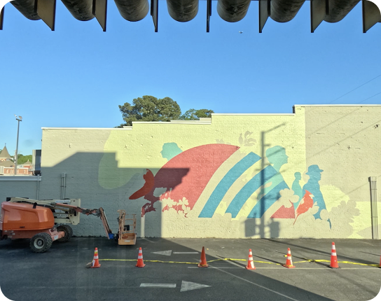 Colorful abstract mural on a pale wall in a parking lot; orange construction lift and orange cones in foreground, under a clear blue sky.