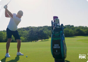 Golfer in a white shirt and cap swings a club on a sunny golf course beside a large golf bag.