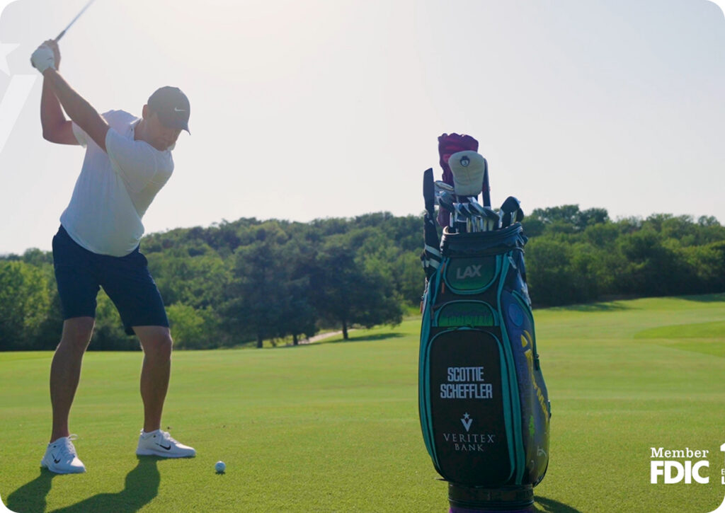 Golfer in a white shirt and cap swings a club on a sunny golf course beside a large golf bag.