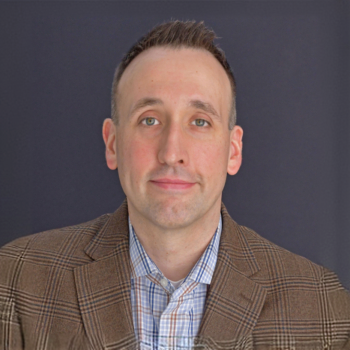 Professional headshot of a man in a brown plaid blazer and blue checkered shirt against a dark blue background.